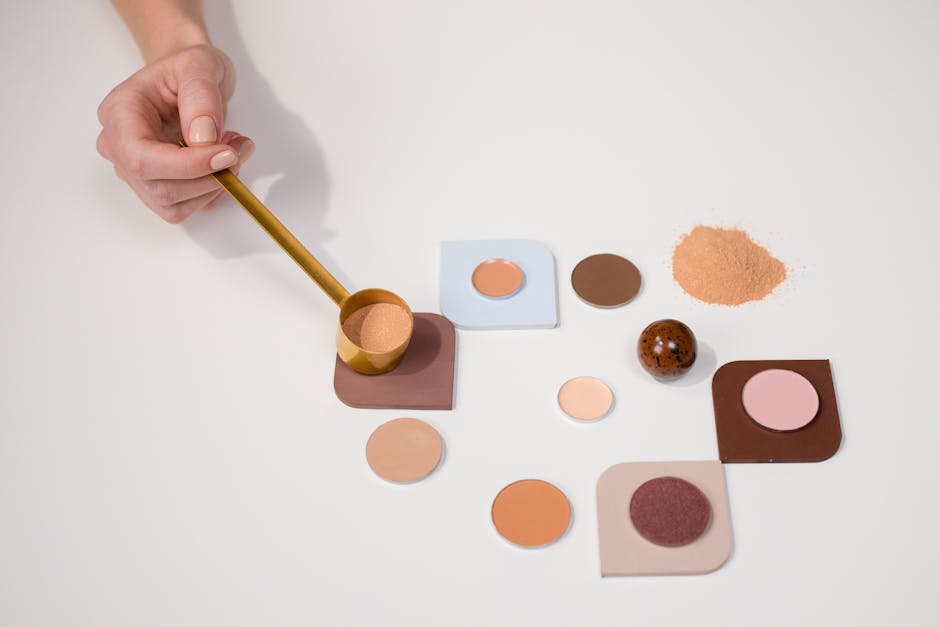 cover-image-276725 Ręka trzymająca złotą miarkę nad różnorodnymi odcieniami kosmetyków do makijażu na jasnym tle. Wśród produktów znajdują się pudry, cienie do powiek oraz drobinki w różnych kolorach.