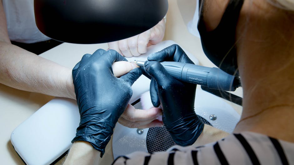 Manicure w salonie kosmetycznym, technik używa narzędzia do pielęgnacji paznokci. W tle widoczna ręka klientki, a nad nimi lampa do manicure.