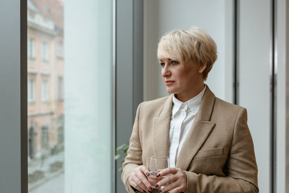Kobieta z krótkimi blond włosami w eleganckim beżowym żakiecie stoi przy dużym oknie, trzymając szklankę. W tle widać miejską architekturę. Obraz przedstawia profesjonalną atmosferę i refleksyjny nastrój.