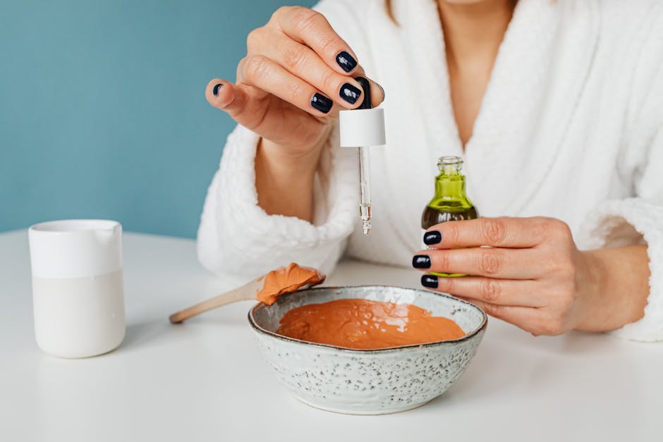 Kobieta w szlafroku trzyma pipetę nad miseczką z brązową maseczką, obok znajduje się butelka z olejkiem i kubek. Idealne dla osób poszukujących inspiracji do pielęgnacji skóry.