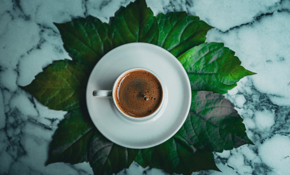Kawa w białej filiżance umieszczona na zielonych liściach, na marmurowym blacie.