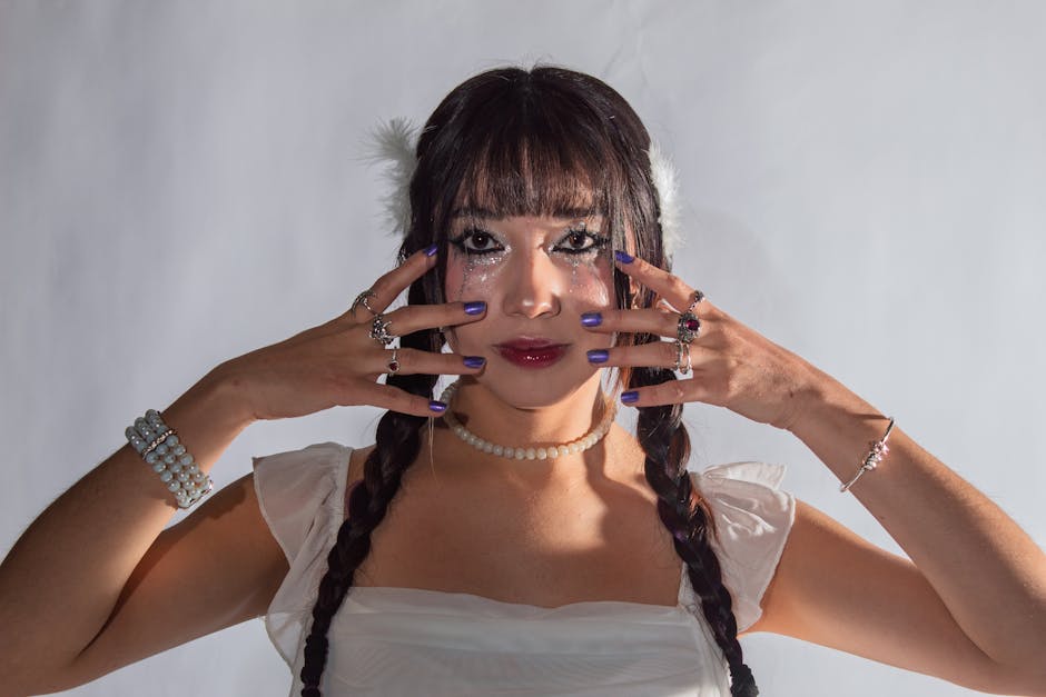 cover-image-276902 Młoda kobieta z długimi warkoczami, ozdobiona biżuterią, prezentuje stylowy makijaż z brokatem. Trzyma dłonie przy twarzy, na paznokciach ma ciemny lakier. Tło jest jasne, co podkreśla jej wyrazisty wygląd.