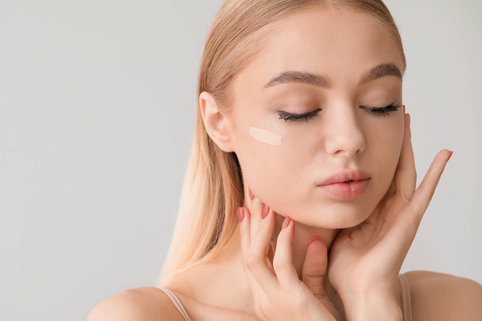 Młoda kobieta z długimi włosami stosująca podkład na twarz, z zamkniętymi oczami, trzymająca dłonie na policzkach, na jasnym tle.